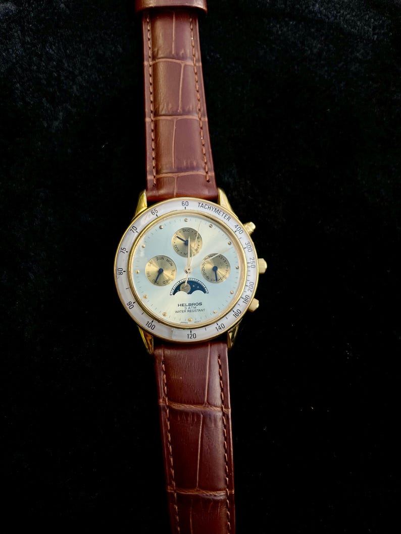 May include: Gold-toned Helbros watch with a light-colored face, featuring multiple subdials and a moon phase indicator. The watch has a brown leather band with a crocodile pattern. The watch face displays the text "TACHYMETER" and "WATER RESISTANT".