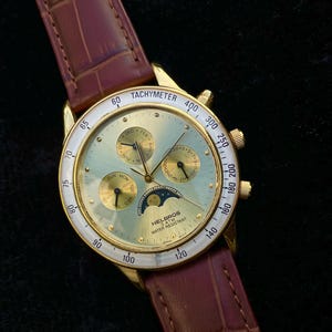 May include: A gold-toned Helbros wristwatch with a light green face, featuring multiple subdials and a tachymeter bezel. The watch has a brown leather band. The watch face displays the text "TACHYMETER" and "WATER RESISTANT".