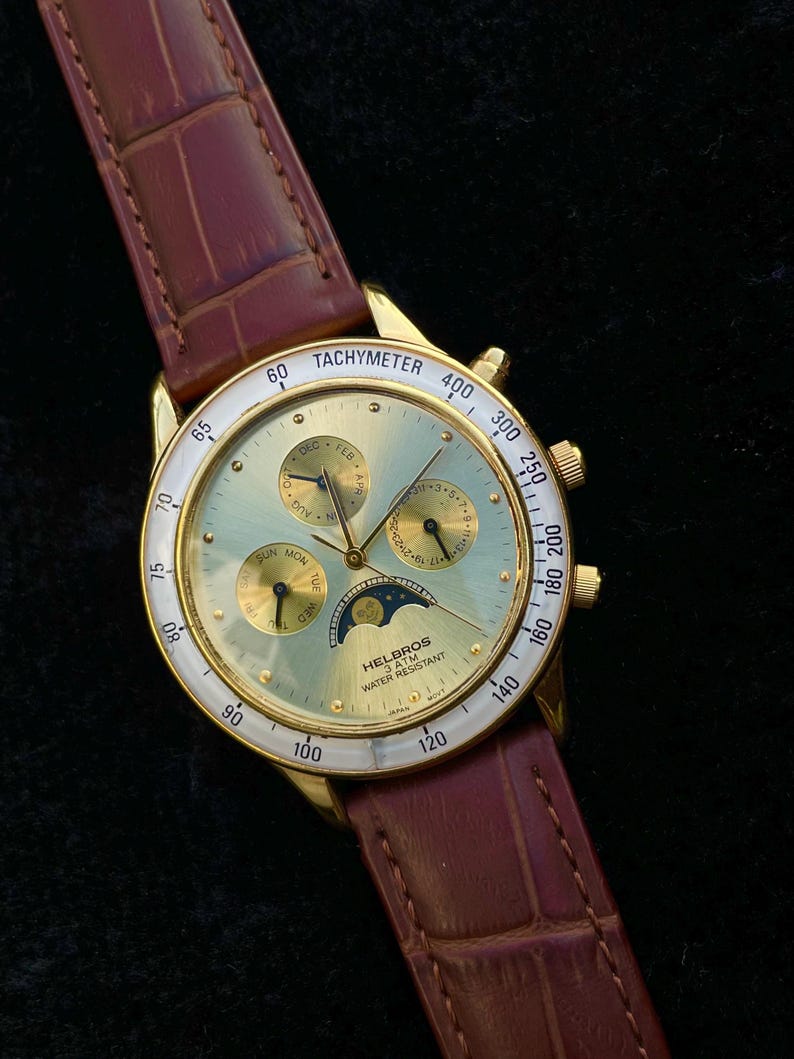 May include: A gold-toned Helbros wristwatch with a light green face, featuring multiple subdials and a tachymeter bezel. The watch has a brown leather band. The watch face displays the text "TACHYMETER" and "WATER RESISTANT".