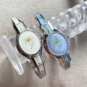 Op de afbeelding: Twee zilverkleurige armbandhorloges met bloemmotieven. Eén horloge heeft een witte wijzerplaat, de andere een lichtblauwe wijzerplaat, beide met een geel en groene bloem. De bandjes zijn versierd met bijpassende bloempatronen.