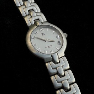 May include: A silver-toned Givenchy watch with a white face and Roman numeral hour markers. The watch features a silver link band and the Givenchy logo. The watch hands point to 10:09. The watch is on a black background.