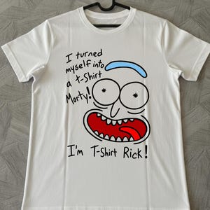 Può includere: Una t-shirt bianca a maniche corte con la grafica del volto di un personaggio dei cartoni animati con occhi grandi, una bocca spalancata con interno rosso e un ciuffo di capelli azzurro. Il testo recita: I turned myself into a t-Shirt Morty! I'm T-Shirt Rick!