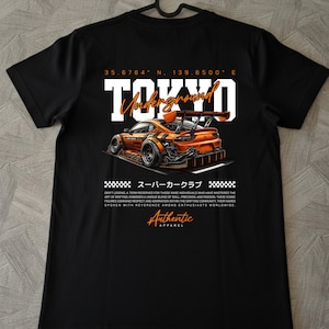 Può includere: T-shirt nero con grafica di un'auto sportiva arancione e la scritta "TOKYO Underground". Il design include testo giapponese e coordinate. La maglietta è appesa a una gruccia nera.