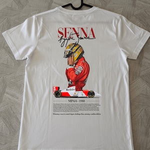 Peut inclure: T-shirt blanc avec un graphisme d'Ayrton Senna en combinaison et casque de course, avec le texte "SENNA" et sa signature. Le design comprend une voiture de course McLaren MP4/4 et le texte "MP4/4 - 1988".