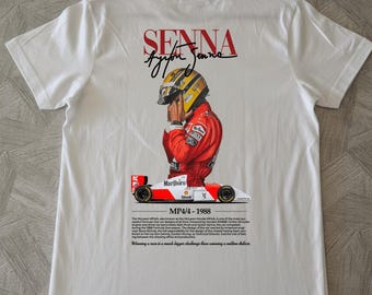 Könnte beinhalten: Ein weißes T-Shirt mit einer Grafik von Ayrton Senna im roten Rennanzug und gelbem Helm über einem McLaren MP4/4 Formel-1-Wagen. Das Design enthält den Text SENNA, MP4/4 - 1988 sowie ein Zitat über das Gewinnen eines Rennens.