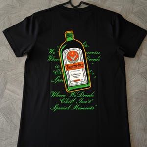 Può includere: Una t-shirt nera a maniche corte con la grafica di una bottiglia di Jägermeister sul retro. La bottiglia ha un contorno verde e arancione. Sotto la bottiglia, un testo in corsivo verde recita: Where We Drink Chill Ion's Special Moments.