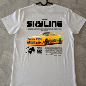 Può includere: Una t-shirt bianca con una stampa grafica sul retro. Il design include il testo NISSAN SKYLINE, un'illustrazione di un'auto sportiva gialla con decalcomanie a fiamma verde, un'icona del globo, loghi di marchi e due blocchi di testo descrittivo.