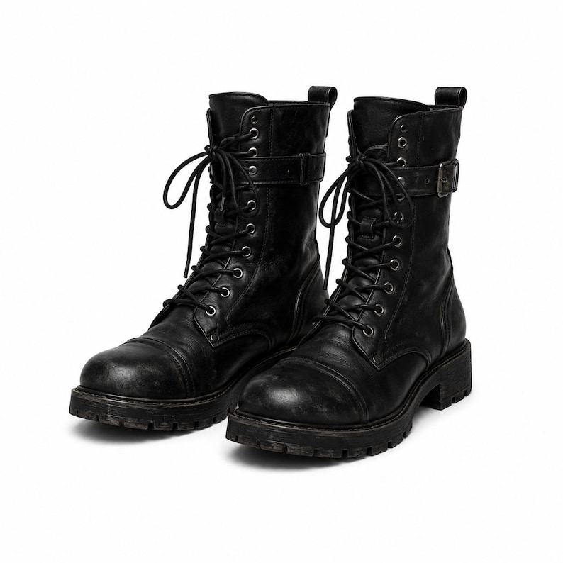 May include: A pair of black mid-calf boots featuring a lace-up front with silver-toned eyelets, a rounded toe cap, and a decorative strap with a metal buckle near the top. The boots have a thick, lugged rubber sole and a pull tab at the heel.