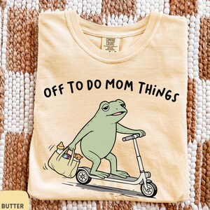 Może przedstawiać: Jasnożółta koszulka z napisem "OFF TO DO MOM THINGS" nad kreskówkową żabą na skuterze, trzymającą torbę z artykułami dla niemowląt. Koszulka jest oznaczona jako "BUTTER".