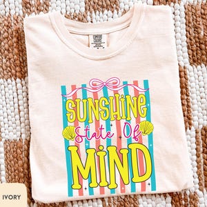 Puede incluir: Camiseta de color marfil con el texto "Sunshine State of Mind" en amarillo, azul y rosa. El diseño incluye conchas marinas y un lazo. La camiseta está hecha de un material suave.