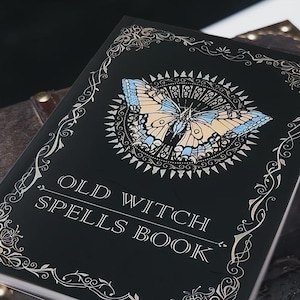 Pu&ograve; includere: Un libro con copertina rigida nera intitolato OLD WITCH SPELLS BOOK in lettere bianche. La copertina presenta un'illustrazione centrale di una farfalla con ali azzurre e beige, circondata da un motivo a raggiera e bordi in filigrana argentata.