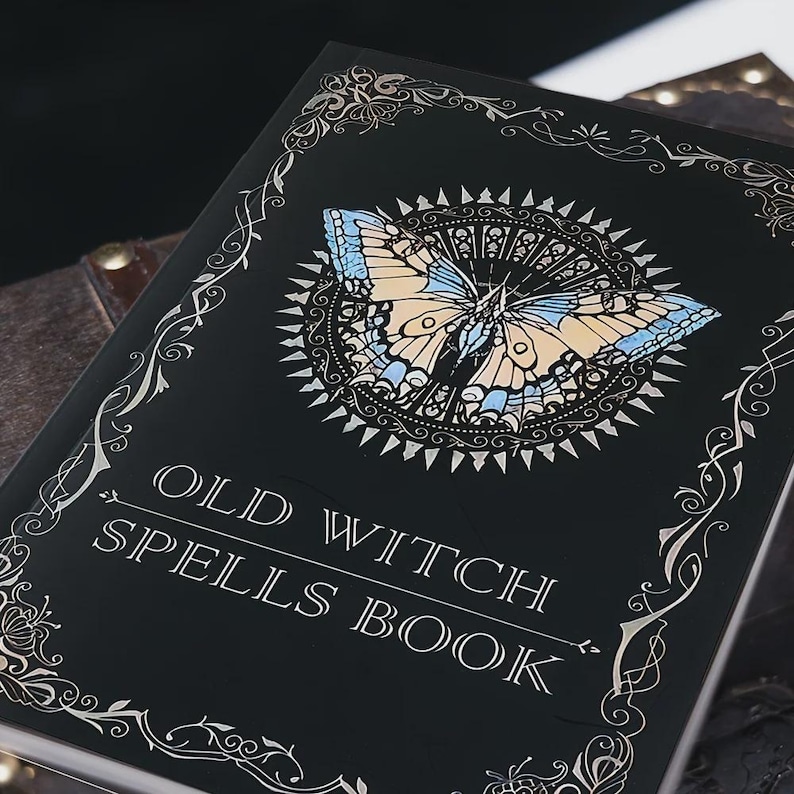Pu&ograve; includere: Un libro con copertina rigida nera intitolato OLD WITCH SPELLS BOOK in lettere bianche. La copertina presenta un'illustrazione centrale di una farfalla con ali azzurre e beige, circondata da un motivo a raggiera e bordi in filigrana argentata.