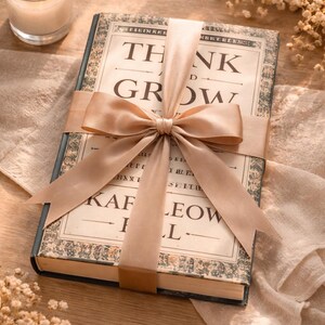 May include: A hardback book titled "THINK AND GROW" wrapped with a beige satin ribbon and bow. The book has an antique design with decorative borders and text. A lit candle and dried flowers are in the background.