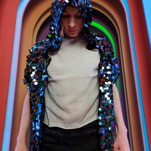 May include: A hooded vest covered in large, iridescent, multi-colored sequins that shift between blue, purple, and green tones. The vest is worn over a white, short-sleeved, mesh-textured top and dark denim pants, creating a layered appearance.