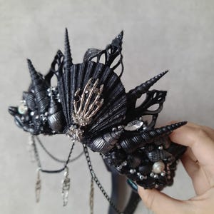 Könnte beinhalten: Eine gotische Krone in Schwarz und Silber, verziert mit Muscheln, Spitzen und hängenden Ketten. Die Krone hat ein fächerförmiges Element mit einer silbernen Skeletthand. Strasssteine und Perlen ergänzen das verzierte Kopfstück.