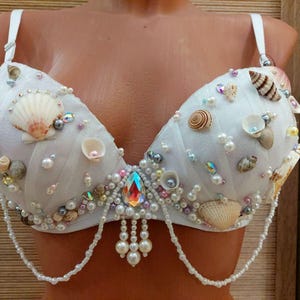 Può includere: Un reggiseno bianco decorato con varie conchiglie, tra cui capesante e conchiglie a spirale, intervallate da perle bianche, perline iridescenti e un cristallo centrale a goccia. Il capo presenta catenelle di perle pendenti sul bordo inferiore.