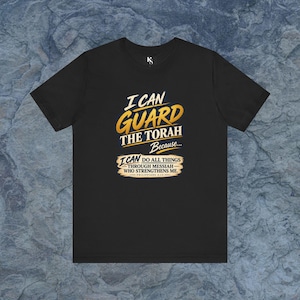 May include: A black short-sleeve t-shirt featuring gold and white text that reads I CAN GUARD THE TORAH Because... I CAN DO ALL THINGS THROUGH MESSIAH WHO STRENGTHENS ME. PHILIPPIANS 4:13. The shirt has a crew neckline and a small white logo at the collar.