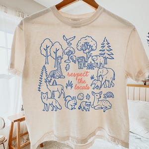 May include: A cream-colored short-sleeve t-shirt featuring a blue graphic print of various forest and wild animals, trees, and foliage. The text "respect the locals" is printed in the center in a coral-colored script font on the front.