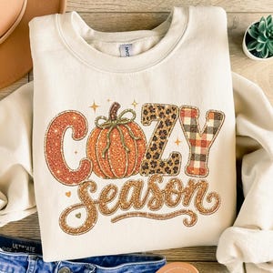 May include: A cream-colored long-sleeve sweatshirt featuring the text Cozy Season in glittery orange, leopard print, and red plaid patterns. A central orange pumpkin graphic with a green stem sits between the letters. The design includes small stars.