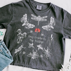 May include: A charcoal gray short-sleeve t-shirt featuring white line art illustrations of various insects, including moths, a dragonfly, a grasshopper, and beetles, with a red ladybug and the text Tiny creatures, Big feelings printed in white.