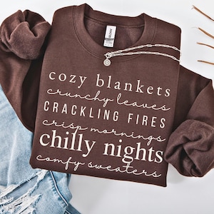 May include: A dark brown crewneck sweatshirt featuring white text that reads: cozy blankets, crunchy leaves, CRACKLING FIRES, crisp mornings, chilly nights, and comfy sweaters. A silver chain necklace rests on the collar of the sweatshirt.