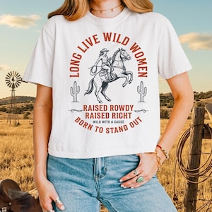 May include: A white short-sleeved t-shirt featuring a graphic of a cowgirl on a horse. The text reads LONG LIVE WILD WOMEN, RAISED ROWDY RAISED RIGHT, and BORN TO STAND OUT. The design includes two cactus illustrations in a rust-colored ink.