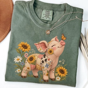 May include: A sage green t-shirt featuring a graphic of a pig with a patchwork pattern of sunflowers, daisies, and gingham checks. The pig has a butterfly on its nose and is surrounded by sunflowers and daisies. The shirt has a Comfort Colors label.