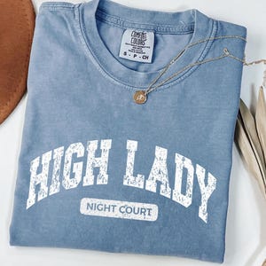 May include: A blue short-sleeved t-shirt with a distressed white graphic print that reads HIGH LADY NIGHT COURT in a collegiate font. The shirt features a Comfort Colors brand label at the inner neckline and is size small.