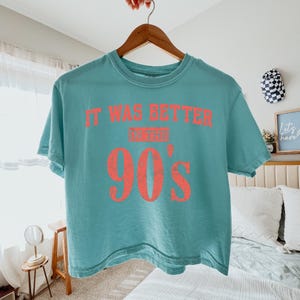 May include: A teal short-sleeved t-shirt featuring the distressed coral-colored text "IT WAS BETTER IN THE 90'S" printed across the chest. The shirt has a crew neckline and is displayed on a wooden hanger against a neutral background.