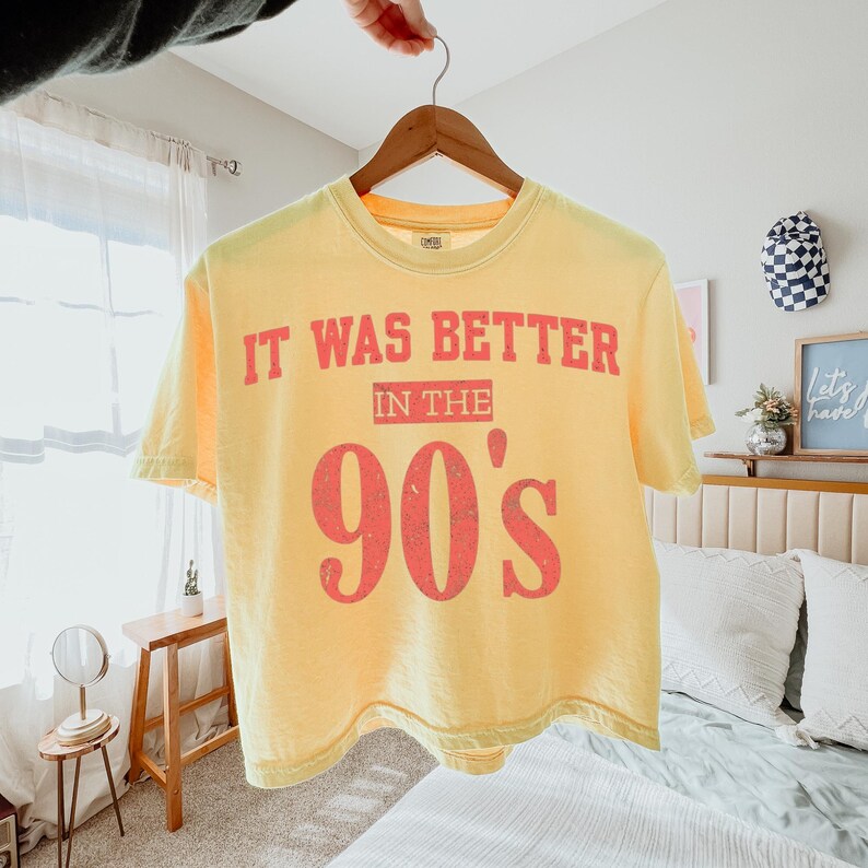 May include: A pale yellow short-sleeve t-shirt featuring the red text "IT WAS BETTER IN THE 90's" printed on the front. The shirt has a crew neckline and a slightly distressed, vintage-inspired graphic design with a weathered texture.