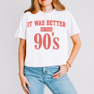 May include: A white short-sleeved t-shirt featuring the red distressed text IT WAS BETTER IN THE 90's printed on the front. The shirt has a crew neckline and a standard fit, paired with blue denim jeans that have a classic five-pocket design.