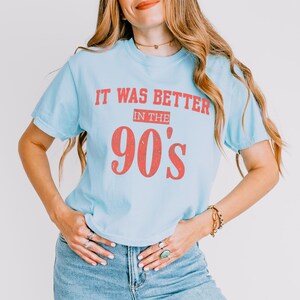 May include: A light blue short-sleeved t-shirt featuring the red text "IT WAS BETTER IN THE 90's" printed on the front. The lettering has a distressed, weathered appearance. The shirt is paired with blue denim jeans, creating a casual look.