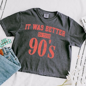 May include: A charcoal gray short-sleeve t-shirt with a distressed red graphic print that reads IT WAS BETTER IN THE 90'S. The shirt features a crew neckline and a vintage-washed fabric texture, displayed alongside blue denim jeans and a small green accessory.