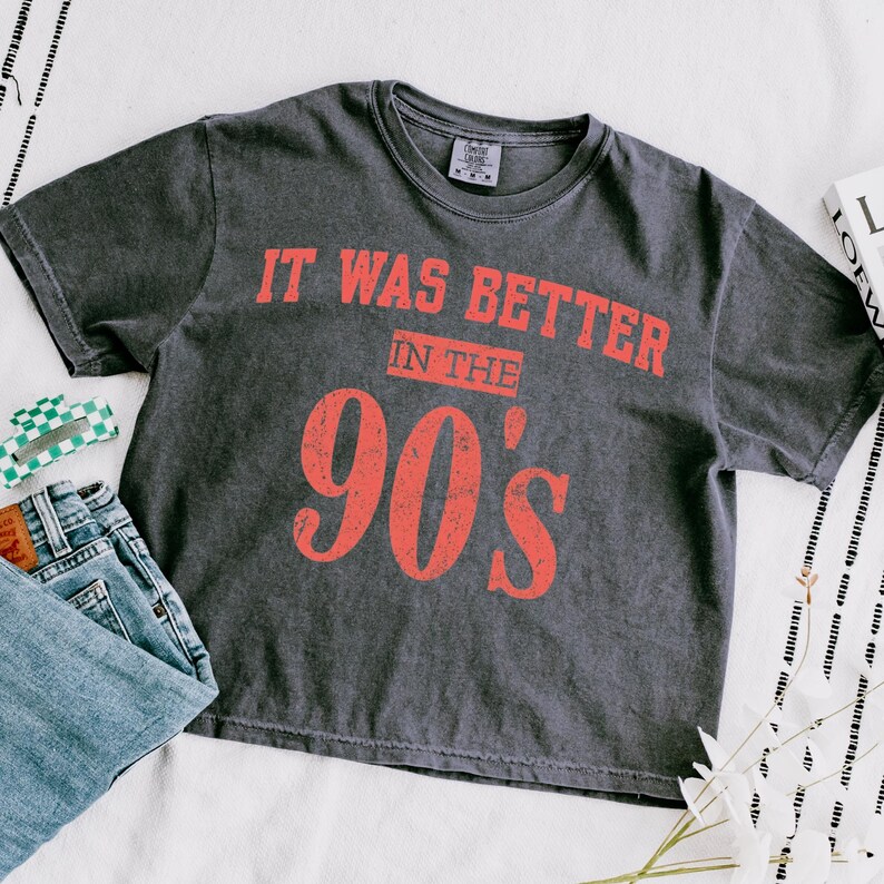 May include: A charcoal gray short-sleeve t-shirt with a distressed red graphic print that reads IT WAS BETTER IN THE 90'S. The shirt features a crew neckline and a vintage-washed fabric texture, displayed alongside blue denim jeans and a small green accessory.