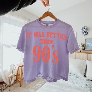 May include: A light purple short-sleeve t-shirt with a crew neckline. The shirt features the orange text "IT WAS BETTER IN THE 90'S" printed across the chest in a distressed, block-style font. The garment is displayed on a wooden hanger.