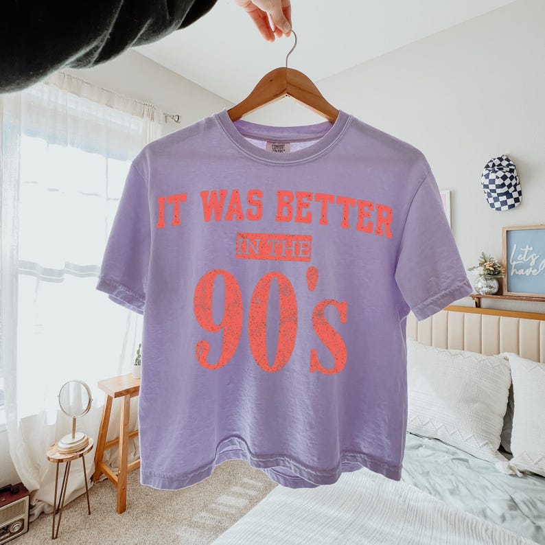May include: A light purple short-sleeve t-shirt with a crew neckline. The shirt features the orange text "IT WAS BETTER IN THE 90'S" printed across the chest in a distressed, block-style font. The garment is displayed on a wooden hanger.