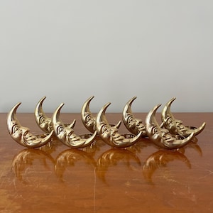 Puede incluir: Un juego de seis servilleteros de metal en tono dorado, cada uno con un diseño de luna creciente con un rostro detallado. Los motivos lunares están dispuestos en fila, mostrando una superficie pulida y reflectante y una forma curva.