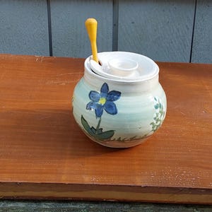 May include: A small, hand-painted ceramic jar with a lid and a yellow spoon. The jar is light blue with a blue flower design and green foliage. The jar is sitting on a wooden surface.