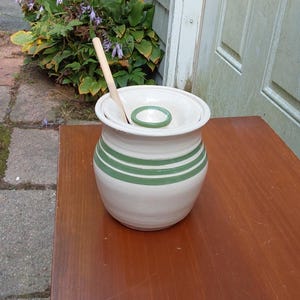 May include: A white ceramic butter dish with three green stripes, a lid, and a small wooden spoon. The dish is sitting on a wooden table. The butter dish is a kitchen item.