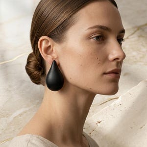 May include: A woman wearing large, black teardrop-shaped earrings. The earrings have a smooth, matte finish, contrasting with the woman's skin. The background is a neutral, textured surface.