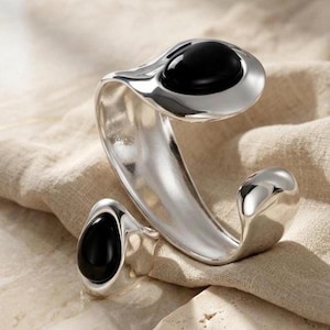 May include: A silver-toned cuff bracelet and ring set. The bracelet and ring feature a smooth, curved design with a polished finish. Each piece has a large, oval-shaped black stone set in the center, creating a striking contrast.