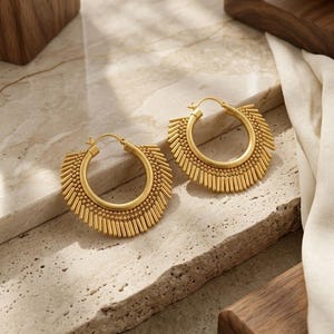 May include: A pair of gold hoop earrings featuring a matte finish. Each earring consists of a circular hoop base adorned with a semi-circular fringe of small vertical metal bars and tiny spherical beads along the outer edge of the hoop.