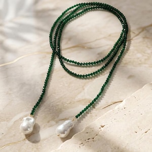 Puede incluir: Un collar largo con múltiples hilos de pequeñas cuentas verdes facetadas. El collar termina con dos grandes perlas blancas de forma irregular. Las perlas están suspendidas de los hilos de cuentas verdes con elementos dorados.