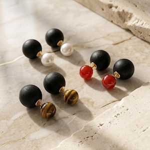 Può includere: Tre paia di orecchini a doppio lato con grandi sfere in legno nero opaco abbinate a sfere più piccole in perla bianca, pietra rossa traslucida e occhio di tigre marrone, collegate da perni in metallo color oro.