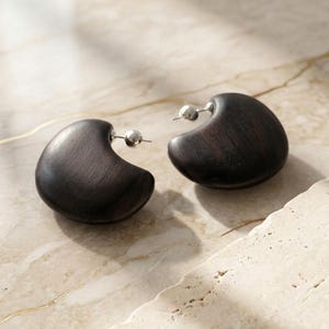 May include: A pair of dark brown, crescent-shaped wooden earrings featuring a smooth, polished finish. Each earring is attached to a small, round, silver-toned metal stud post, providing a contrast between the natural wood grain and the metallic hardware.