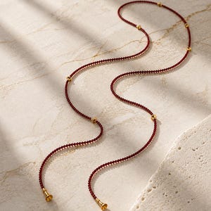 May include: A long, delicate necklace composed of tiny, deep red beads arranged in a continuous strand. The necklace is accented with small, polished gold-toned metal beads spaced at intervals and features two gold-toned bell-shaped end caps.