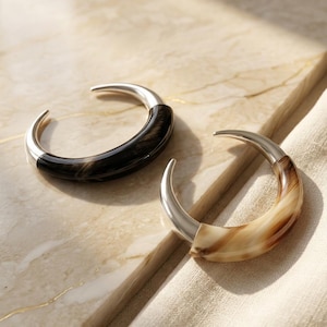 May include: Two crescent-shaped bracelets with silver-toned metal accents. One bracelet is black and the other is tan and brown. The bracelets are displayed on a light-colored surface.
