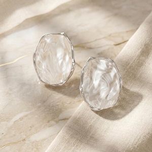 May include: Two silver-toned, irregular-shaped earrings with a pearlescent, swirled interior. The earrings are set against a neutral-toned background, with a soft, textured fabric and a marble-like surface.