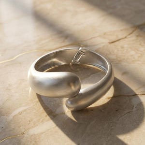 Puede incluir: Un brazalete de puño curvo de color plateado con cierre de bisagra. El brazalete tiene un acabado liso y mate y un diseño redondeado. La joya se exhibe sobre una superficie clara, similar al mármol, con sombras.