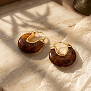 May include: Two hoop earrings with a tortoiseshell pattern. The earrings feature a gold-coloured metal clasp and a rounded, semi-circular design. The brown and amber hues of the earrings are set against a neutral backdrop.